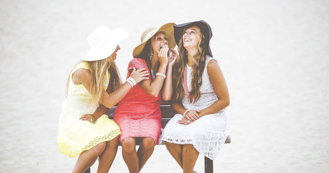 Auf dem Bild sieht man drei junge Frauen auf einer Bank sitzen, die mit einander lachen. Dabei stecken sie ihre Köpfe zusammen. Die Frau links trägt ein gelbes, die Frau in der Mitte ein rotes und die Frau rechts ein weißes Sommerkleid.