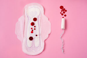 Auf der Abbildung ist eine Damenbinde und ein Tampon auf einem rosa Hintergrund zu sehen. Blutstropfen sind durch rote Steine dargestellt. 
