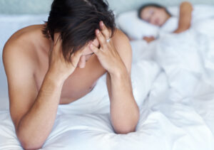Junger Mann sitzt aufrecht im Bett und hält seine Hände frustrierend vor die Stirn, während seine Frau im Hintergrund schläft.