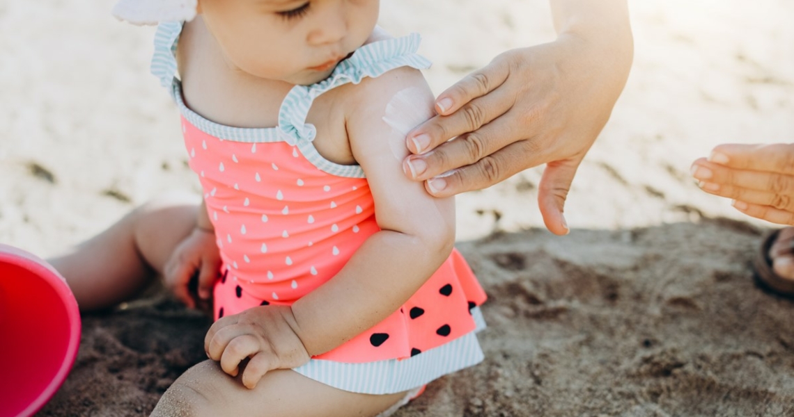 Das Bild zeigt ein kleines Kind, welches im Sand spielt. Eine weibliche Hand cremt den Oberarm des Kindes mit Sonnencreme ein.
