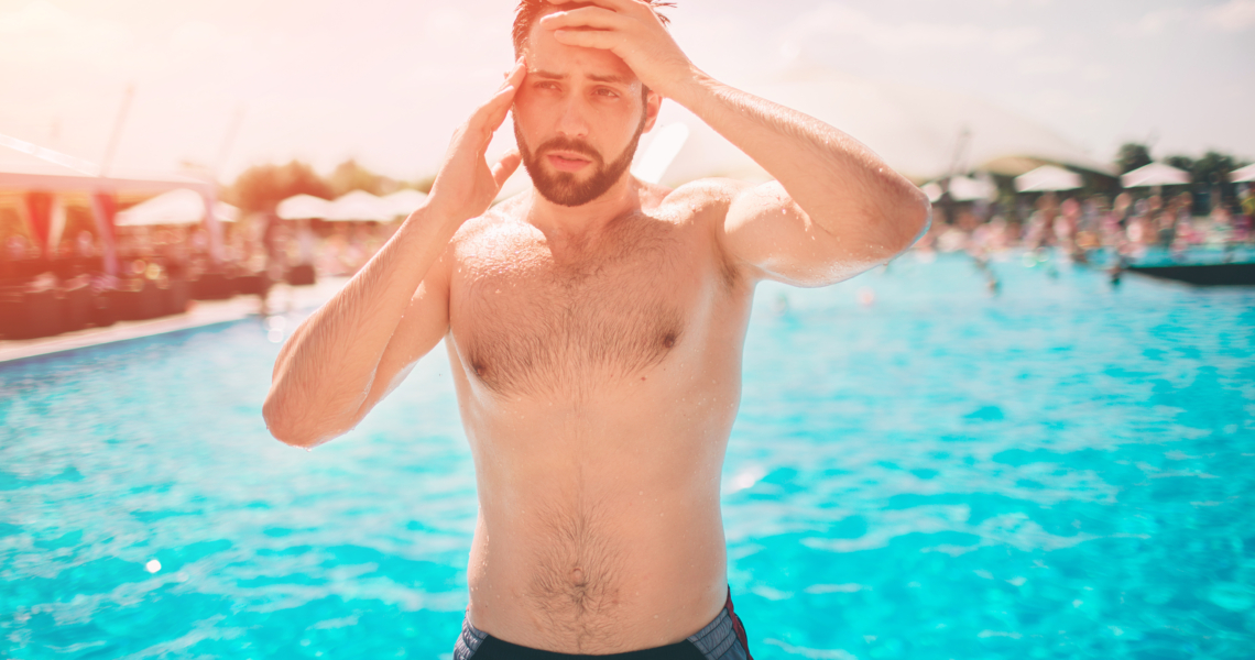 Das Bild zeigt einen Mann, der von einem Pool in Badeshorts steht und sich mit beiden Händen an den Kopf fässt.
