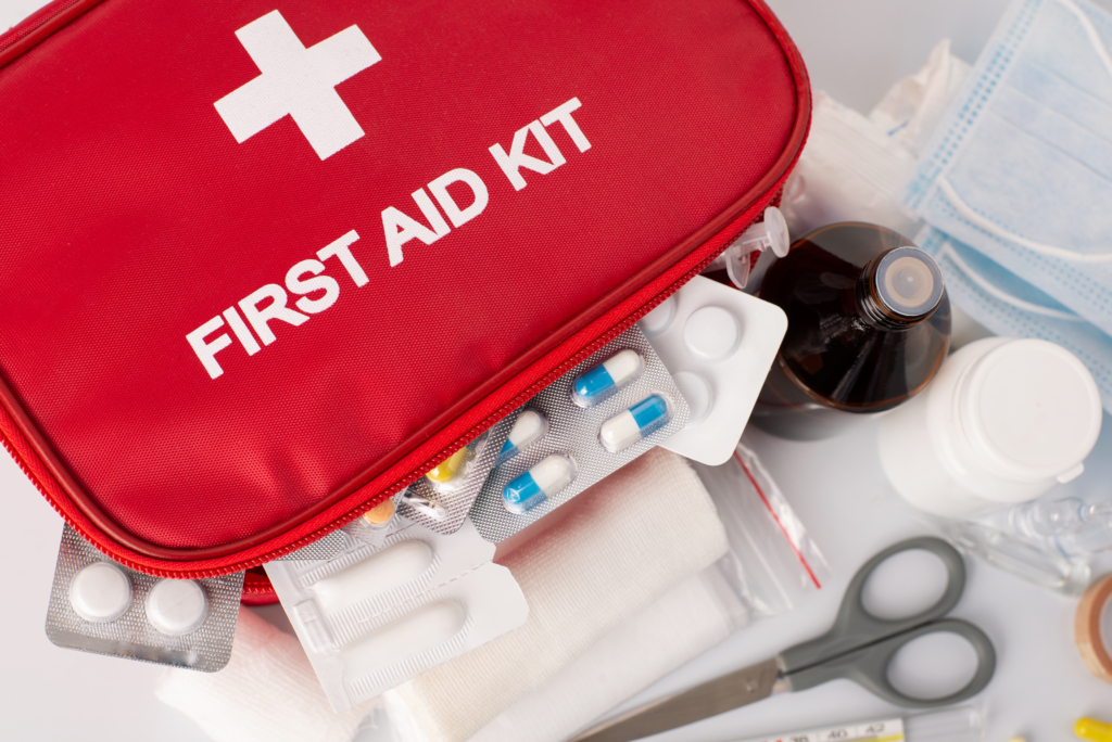 Das Bild zeigt eine rote Medikamententasche mit einem weißen Kreuz und der Aufschrift "First Aid Kit". Mehrere Medikamentenblister befinden sich in der Tasche sowie außerhalb der Tasche.