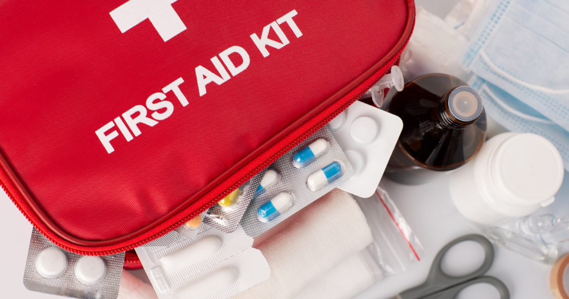 Das Bild zeigt eine rote Medikamententasche mit einem weißen Kreuz und der Aufschrift "First Aid Kit". Mehrere Medikamentenblister befinden sich in der Tasche sowie außerhalb der Tasche.