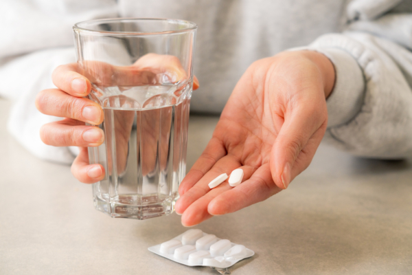 Das Bild zeigt eine Person, die in der einen Hand ein Glas und in der anderen zwei Ibuprofen Tabletten hat. Vor ihr liegt ein Blister mit Ibuprofen.