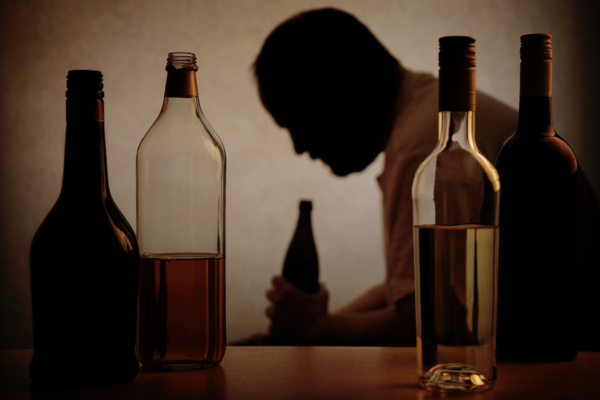 Das Bild zeigt Alkoholflaschen im Vordergrund, im Hintergrund sieht man die Silhouette eines Mannes mit einer Bierflasche in der Hand. 