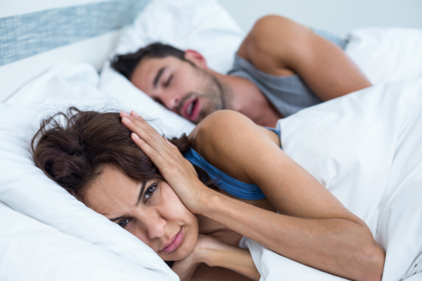 Das Bild zeigt eine Frau und einen Mann im Bett. Der Mann schnarcht und schläft, die Frau ist wach und hält sich die Ohren zu. 