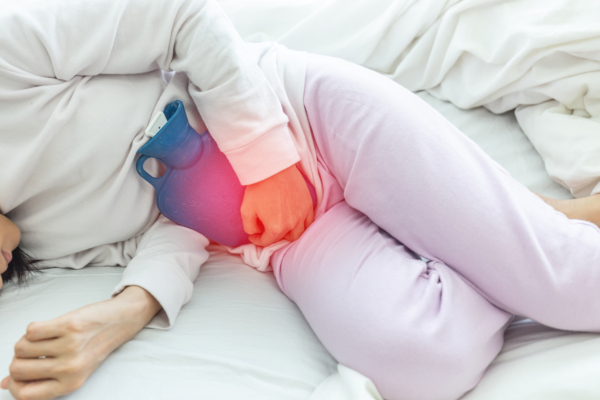 Das Bild zeigt eine Frau, die gekrümmt im Bett liegt und eine Wärmflasche auf dem Bauch hat. 
