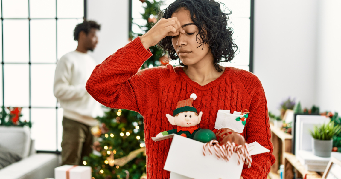 Eine Frau steht gestresst mit Geschenken in der Hand vor einem Weihnachtsbaum