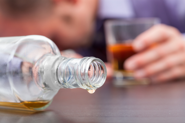 Das Bild zeigt eine leere Schnapsflasche, die auf dem Tisch liegt, im Hintergrund liegt ein Mann mit dem Kopf auf dem Tisch und hält in der Hand noch ein Glas voller Alkohol. 