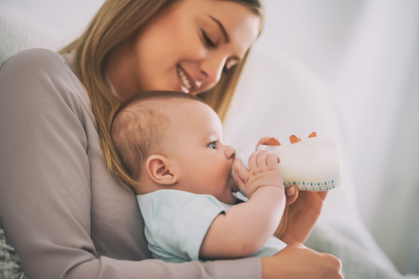 Das Bild zeigt eine Mutter, die ihr Baby auf dem Arm hält, welches an einer Nuckelflasche nuckelt. 