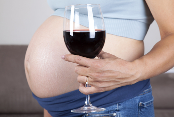 Das Bild zeigt eine schwangere Frau, die ein Glas Rotwein in der Hand hält. 