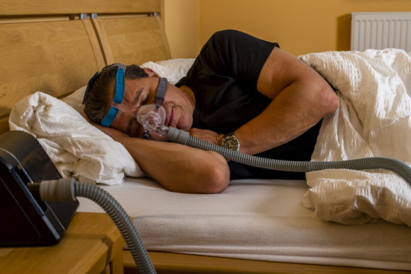 Das Bild zeigt einen schlafenden Mann im Bett, der ein Beatmungsgerät trägt.