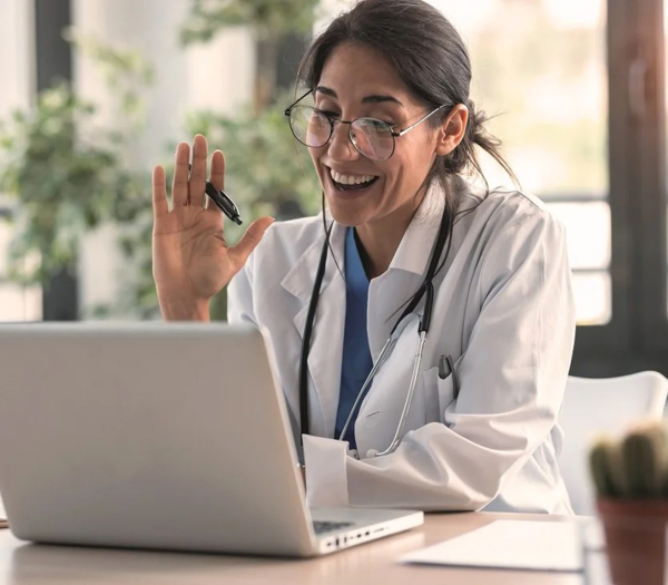 Ärztin mit Stethoskop führt lächelnd eine Videosprechstunde am Laptop durch und winkt in die Kamera.